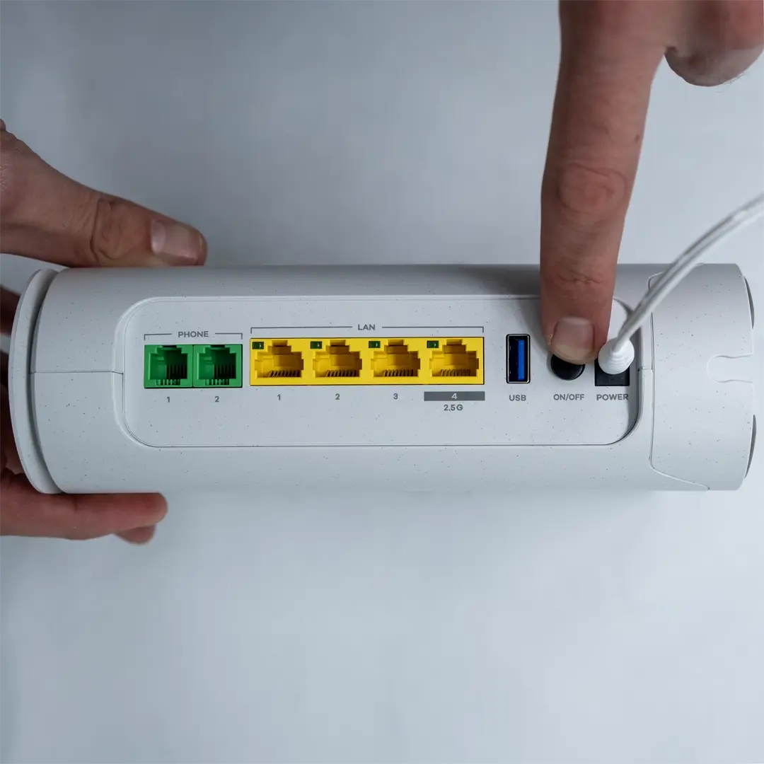 Eine Person drückt die Einschalttaste auf der Rückseite eines Zyxel-Routers, um ihn zu starten. Das Netzkabel ist angeschlossen, und die gelben LAN-Ports sowie der USB-Port sind deutlich sichtbar.