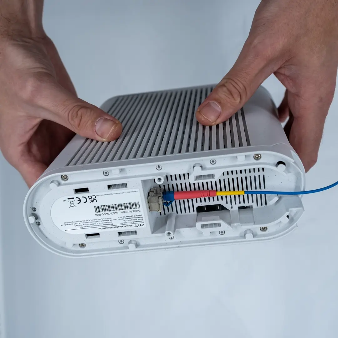 Eine Person hält den Router umgedreht und zeigt, wie das SFP-Modul mit angeschlossener blauer Glasfaserkabel in den Anschluss an der Unterseite eingesetzt wird.