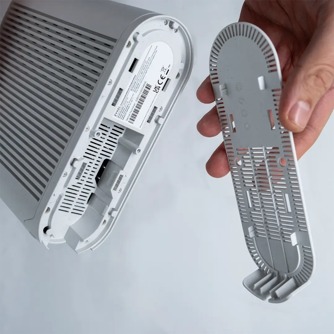 Eine Person hat die Bodenabdeckung vom Router entfernt und hält sie neben das Gerät, sodass die Anschlüsse an der Unterseite sichtbar sind.